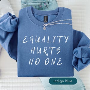 May include: Indigo blue sweatshirt with the text "EQUALITY HURTS NO ONE" in white. A silver necklace with a pendant is draped across the sweatshirt. White sneakers and blue jeans are visible in the background.