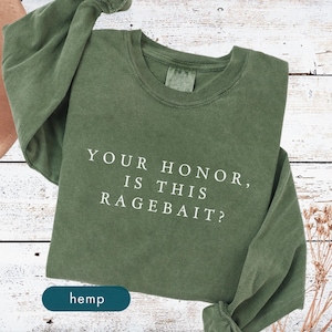 May include: Olive green long-sleeve shirt with the text "YOUR HONOR, IS THIS RAGEBAIT?" in white. The word "hemp" is printed on a dark teal tag. The shirt is made of a soft material.