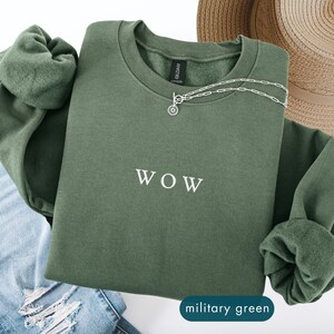 May include: A military green sweatshirt with the word "WOW" in white lettering. A silver necklace with a circular pendant is on the sweatshirt. A straw hat and blue jeans are also in the image.