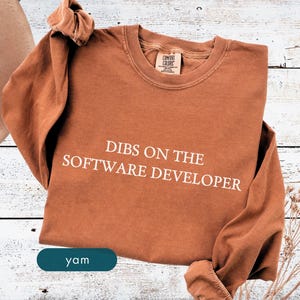 May include: A long-sleeved, rust-colored shirt with the phrase "DIBS ON THE SOFTWARE DEVELOPER" printed in white. The shirt has a crew neck and a tag that reads "COMFORT COLORS". The word "yam" is on a teal colored button.