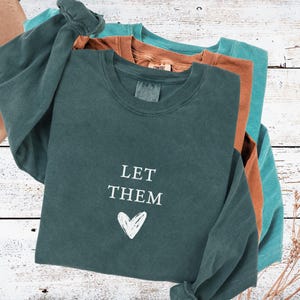 May include: A stack of long-sleeved shirts in teal, rust, and dark teal. The front shirt has the words "LET THEM" and a heart graphic in white. The shirts are on a white wooden surface.