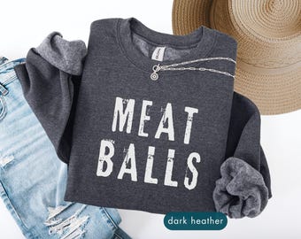 May include: Dark heather gray sweatshirt with the text "MEAT BALLS" in distressed white lettering. A silver necklace and a straw hat are also visible. The sweatshirt is folded with the sleeves tucked in.