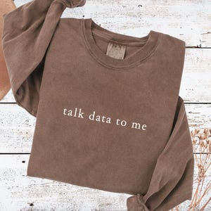 May include: A brown crewneck sweatshirt featuring the text "talk data to me" printed in white across the chest. The garment has long sleeves, a ribbed collar, and a relaxed fit, showcasing a solid, muted brown color throughout the fabric.