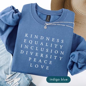 May include: Indigo blue sweatshirt with the words KINDNESS, EQUALITY, INCLUSION, DIVERSITY, PEACE, and LOVE in white block letters. A silver necklace with a small pendant rests on the shirt. Light blue jeans and a straw hat are also in the image.