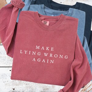 May include: A stack of long-sleeved shirts in shades of red, blue, and navy. The top shirt is a dusty rose color with the text "MAKE LYING WRONG AGAIN" in white. The shirts are folded on a light wood surface.