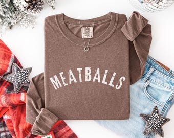 May include: A brown long-sleeve shirt with the word "MEATBALLS" in white, arched across the front. The shirt is folded with a silver necklace and a pair of blue jeans. Decorative stars and a plaid scarf are also present.