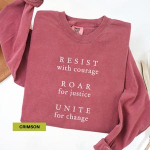 May include: Crimson long-sleeve shirt with the words "RESIST with courage, ROAR for justice, UNITE for change" in white. The shirt is a solid crimson color and has a crew neck.