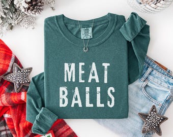 May include: Teal long-sleeve shirt with the words "MEAT BALLS" in white block letters. A silver necklace with a sun pendant is visible. The shirt is paired with blue jeans and decorative stars.