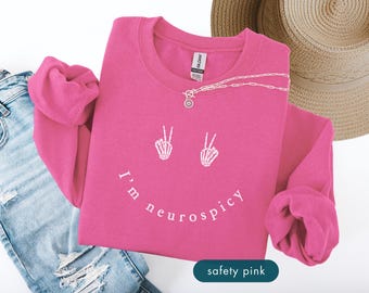 May include: Safety pink sweatshirt with white text and skeleton hand peace signs forming a smile. The text reads "I'm neurospicy". A silver necklace and a straw hat are also visible.