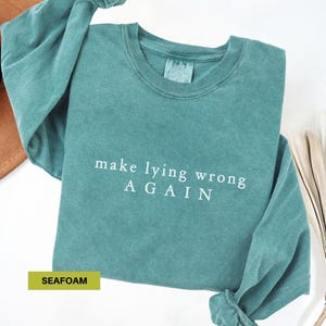 May include: Seafoam green long-sleeved sweatshirt with the phrase "make lying wrong AGAIN" in white block letters. The sweatshirt is made of a soft material and has a crew neck. The word "SEAFOAM" is in a yellow box.