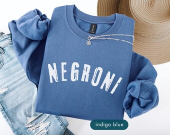 May include: Indigo blue sweatshirt with the word "NEGRONI" printed in white. A silver necklace with a small pendant rests on the sweatshirt. Light blue denim jeans and a straw hat are also in the image.