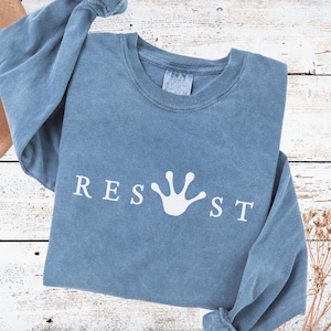 May include: A blue long-sleeve shirt with the word "RESIST" printed in white, featuring a white hand graphic. The shirt is made of a soft material and is displayed on a white wooden surface.