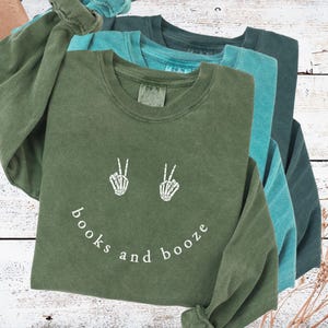 May include: Three long-sleeved shirts in shades of green and teal. The front shirt is olive green with a white graphic of skeleton hands making peace signs and the words "books and booze". The shirts are folded on a white wooden surface.