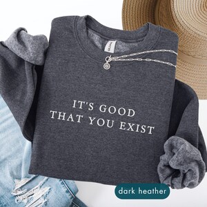 May include: Dark heather gray sweatshirt with the text "IT'S GOOD THAT YOU EXIST" in white. A silver necklace and a straw hat are also in the image. The sweatshirt is folded with the sleeves showing.