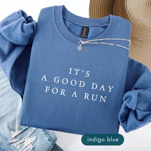 May include: Indigo blue sweatshirt with the text "IT'S A GOOD DAY FOR A RUN" in white. The sweatshirt is paired with light blue jeans, a straw hat, and a silver necklace.