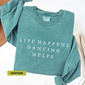 May include: Seafoam green sweatshirt with the text "LIFE HAPPENS DANCING HELPS" in white. The long-sleeved shirt is made of a soft material and has a crew neck. The shirt is folded on a white surface.