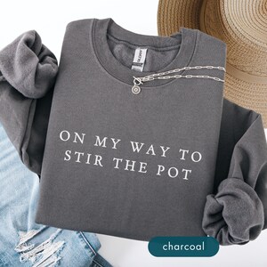 May include: Charcoal gray sweatshirt with the text "ON MY WAY TO STIR THE POT" in white. A silver necklace with a pendant is draped across the front. A straw hat and blue jeans are also in the image.