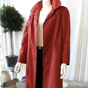 Puede incluir: Un abrigo largo de color óxido con un cuello ancho y cierre de botones. El abrigo se exhibe en un maniquí, combinado con pantalones negros y una parte superior blanca. Se ve un collar de perlas. El abrigo tiene un diseño clásico y entallado.