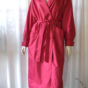 May include: A vibrant pink trench coat with a double-breasted design, long sleeves, and a matching belt. The coat has a classic collar, button details, and pockets. The coat is displayed on a mannequin.