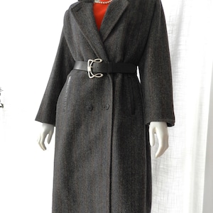 May include: A long, dark gray herringbone coat with a black leather collar and belt. The coat is double-breasted and has a silver buckle on the belt. The mannequin is wearing a red top and a pearl necklace.