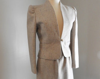 May include: A gray tweed skirt suit with a jacket featuring a V-neck and puffed shoulders. The jacket has a single button closure and a small pocket on the front.