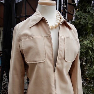 May include: A beige zip-up jacket with a collar and two large pockets on the front. The jacket is on a mannequin wearing a pearl necklace.
