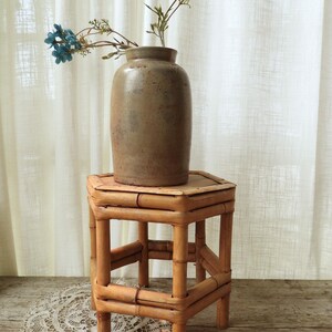 May include: A tall, cylindrical, light brown stoneware crock with a smooth finish sits atop a small, square bamboo stool. A single stem with several small, bright blue flowers extends from the opening of the crock, adding a touch of color to the neutral-toned ceramic vessel and the natural bamboo structure.