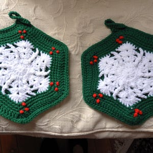 May include: Two handmade, green and white crochet potholders. Each has a hexagonal shape with a white snowflake design in the center, surrounded by a green border. Small red accents are placed around the edges. A loop is attached for hanging.