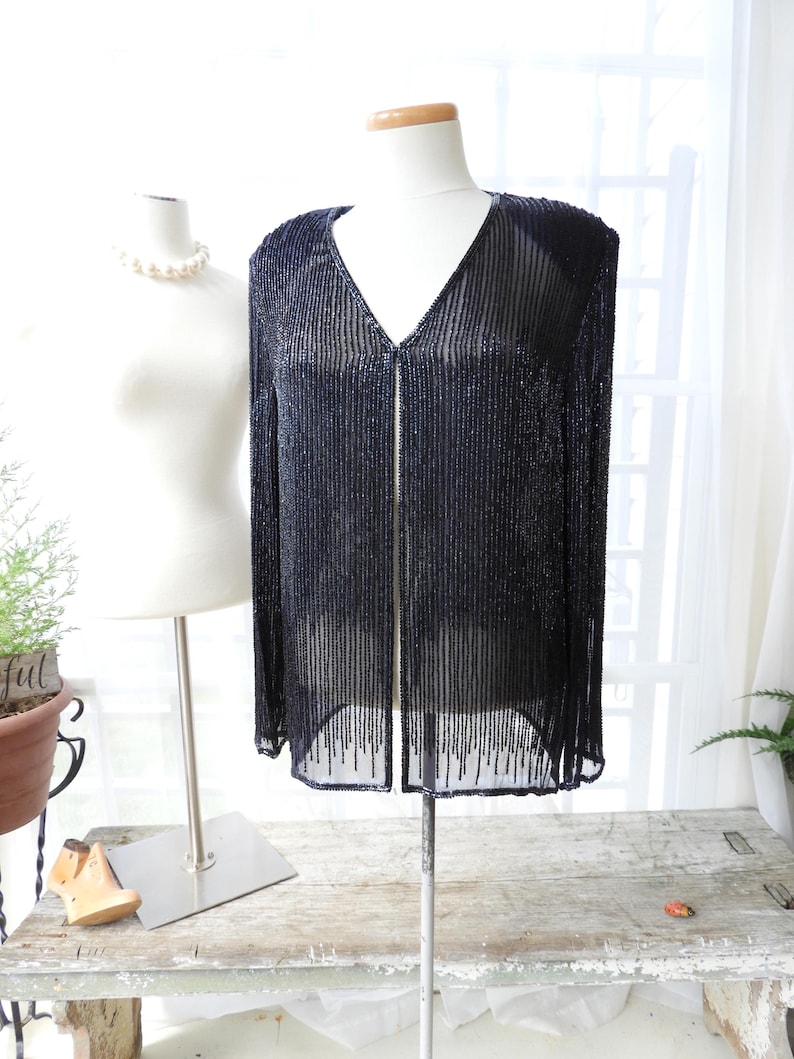 May include: A black beaded cardigan with a V-neckline and long sleeves. The cardigan is made of a sheer fabric and has a beaded fringe at the hem.