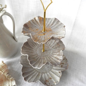 Op de afbeelding: Een serveerschaal met drie niveaus en een gouden handvat. De schalen zijn gevormd als gestileerde bloemen met een getextureerde, grijze afwerking en gouden accenten. De schaal is ontworpen voor het presenteren van voedsel of decoratieve items.