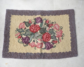 Puede incluir: Alfombra rectangular con diseño floral. La alfombra presenta un ramo central de flores rojas, rosas y moradas con hojas verdes, sobre un fondo beige. El borde es un borde festoneado en un morado apagado.