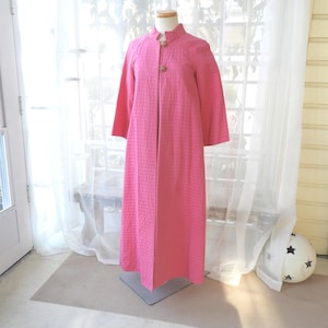 May include: A long, pink, quilted robe with a mandarin collar and three-quarter sleeves. The robe has two decorative buttons at the neckline and is displayed on a mannequin. The fabric has a textured, grid-like pattern.