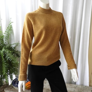May include: Mustard-coloured jumper with a mock neck and long sleeves. The cuffs and neckline are ribbed. The jumper is paired with black trousers. The mannequin is in front of a white curtain.