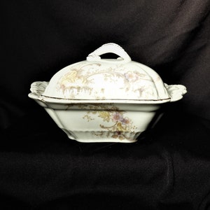 May include: A white porcelain serving dish with a lid. The dish has a floral pattern in shades of pink, yellow, and brown. The lid has a knob on top.