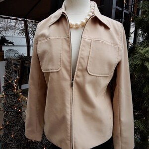 May include: A beige zip-up jacket with two flap pockets on the front. The jacket is on a white mannequin and has a pearl necklace around the neck.
