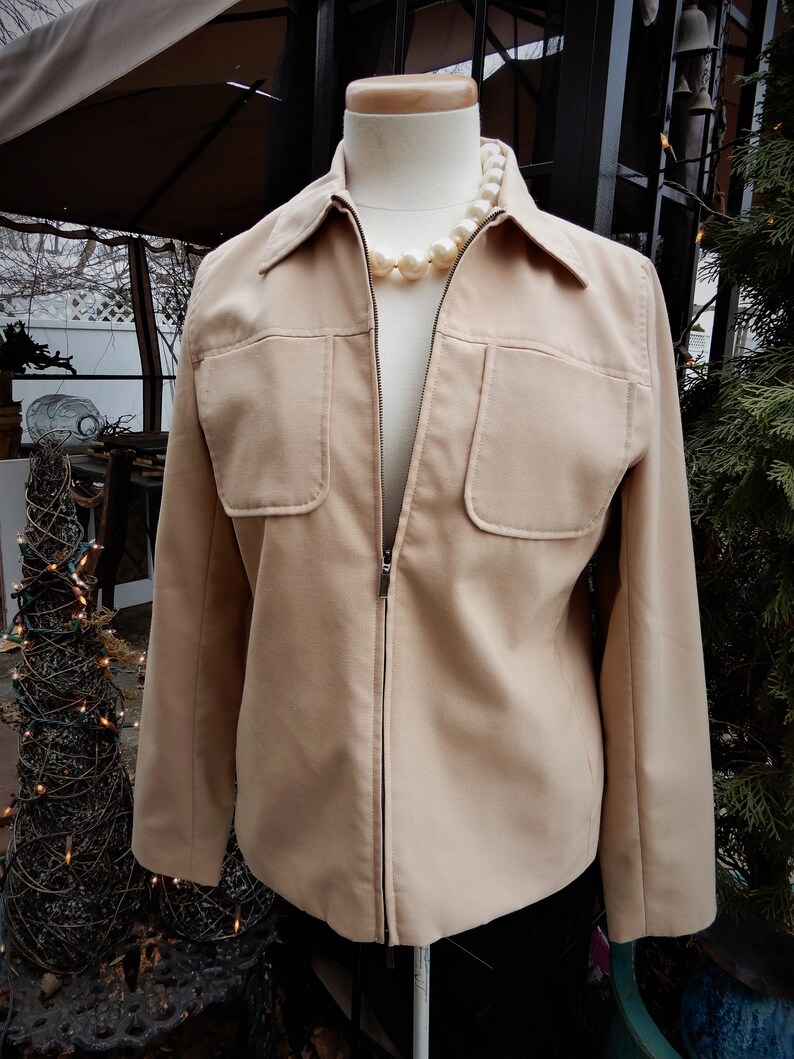 May include: A beige zip-up jacket with two flap pockets on the front. The jacket is on a white mannequin and has a pearl necklace around the neck.