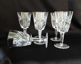 May include: Five clear crystal wine glasses with intricate cut patterns. The glasses have a stemmed base and a detailed design on the bowl. One glass is lying on its side, while the others stand upright against a black background.