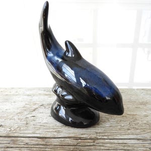 Op de afbeelding: Een zwart keramiek dolfijnbeeldje met een blauwe glazuur. De dolfijn springt uit het water met zijn staart in de lucht.