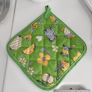 May include: A square, quilted pot holder in a vibrant green color featuring a repeating pattern of yellow flowers, white daisies, sliced citrus fruits, strawberries, small hearts, grey coffee mugs, and coffee grinders, finished with a green fabric loop.
