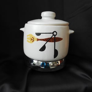 Op de afbeelding: Een witte keramische ovenschaal met deksel, met een retro ontwerp van een gele vork, bruine lepel en zwarte keukengerei. De schaal staat op een verchroomde basis. De achtergrond is zwart.