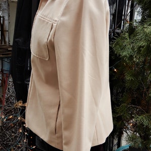May include: A beige blazer with a long sleeve and a single pocket on the left side. The blazer is on a mannequin and is in front of a black metal structure.