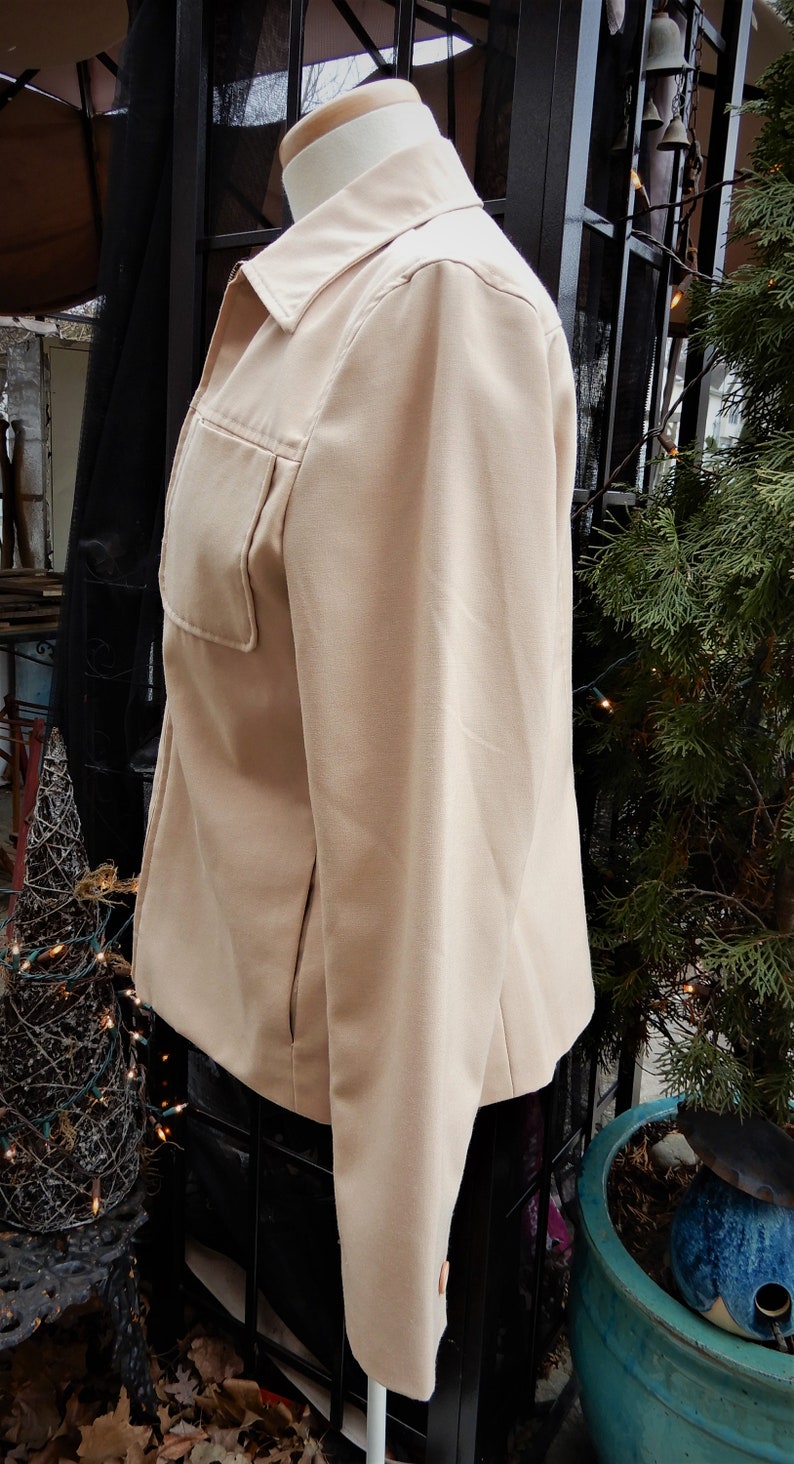 May include: A beige blazer with a long sleeve and a single pocket on the left side. The blazer is on a mannequin and is in front of a black metal structure.