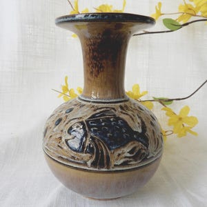 May include: A ceramic vase with a bulbous base and a flared top. The vase is primarily brown with a darker brown interior and a textured design featuring a fish. The vase is approximately 8 inches tall.