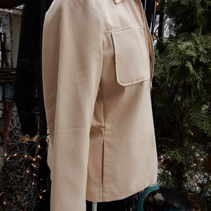 May include: A beige zip-up jacket with a large pocket on the left side. The jacket has a collar and long sleeves.