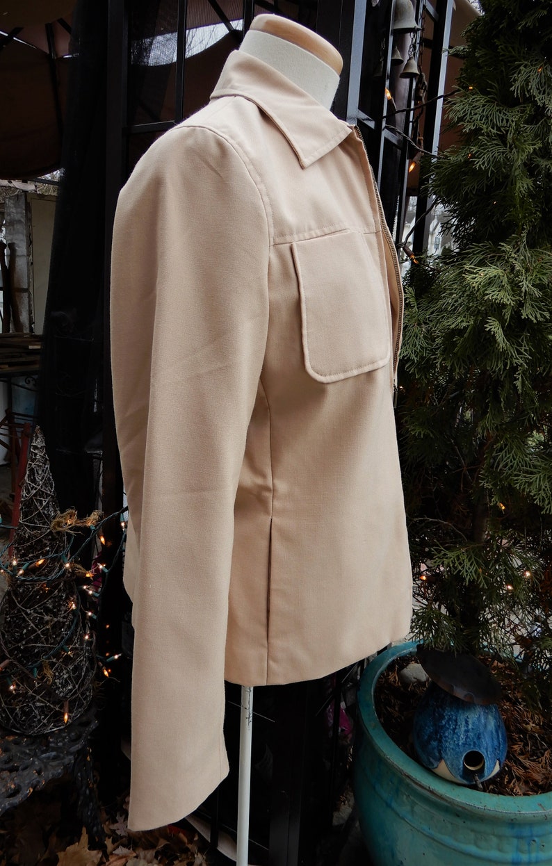 May include: A beige zip-up jacket with a large pocket on the left side. The jacket has a collar and long sleeves.