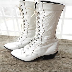 May include: A pair of white leather lace-up boots with a pointed toe and a low heel. The boots have a decorative stitching detail on the side and a tassel on the front.