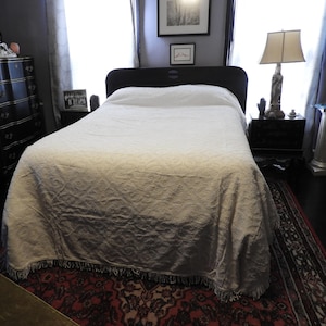 May include: A bedroom featuring a bed with a white, textured bedspread and a dark wooden headboard. A patterned rug in shades of red and black is on the floor. A black dresser and a lamp with a decorative base are also visible.