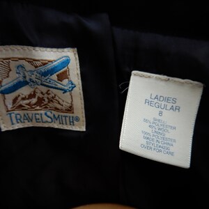 May include: A white tag with black text that reads "LADIES REGULAR 8 SHELL 55% POLYESTER 45% WOOL 100% POLYESTER LINING MADE IN CHINA STYLE#4230 OVER FOR CARE" attached to a black garment with a brown and white logo that reads "TRAVELSMITH".