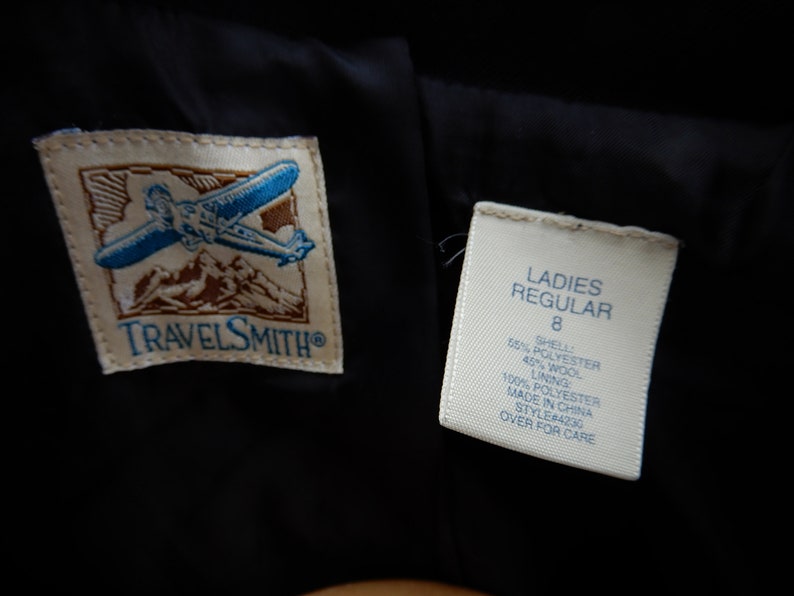 May include: A white tag with black text that reads "LADIES REGULAR 8 SHELL 55% POLYESTER 45% WOOL 100% POLYESTER LINING MADE IN CHINA STYLE#4230 OVER FOR CARE" attached to a black garment with a brown and white logo that reads "TRAVELSMITH".