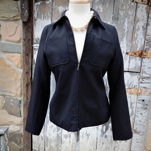 May include: A black zippered jacket with a collared neckline and two flap pockets on the front. The jacket is on a white mannequin and has a pearl necklace around the neck.